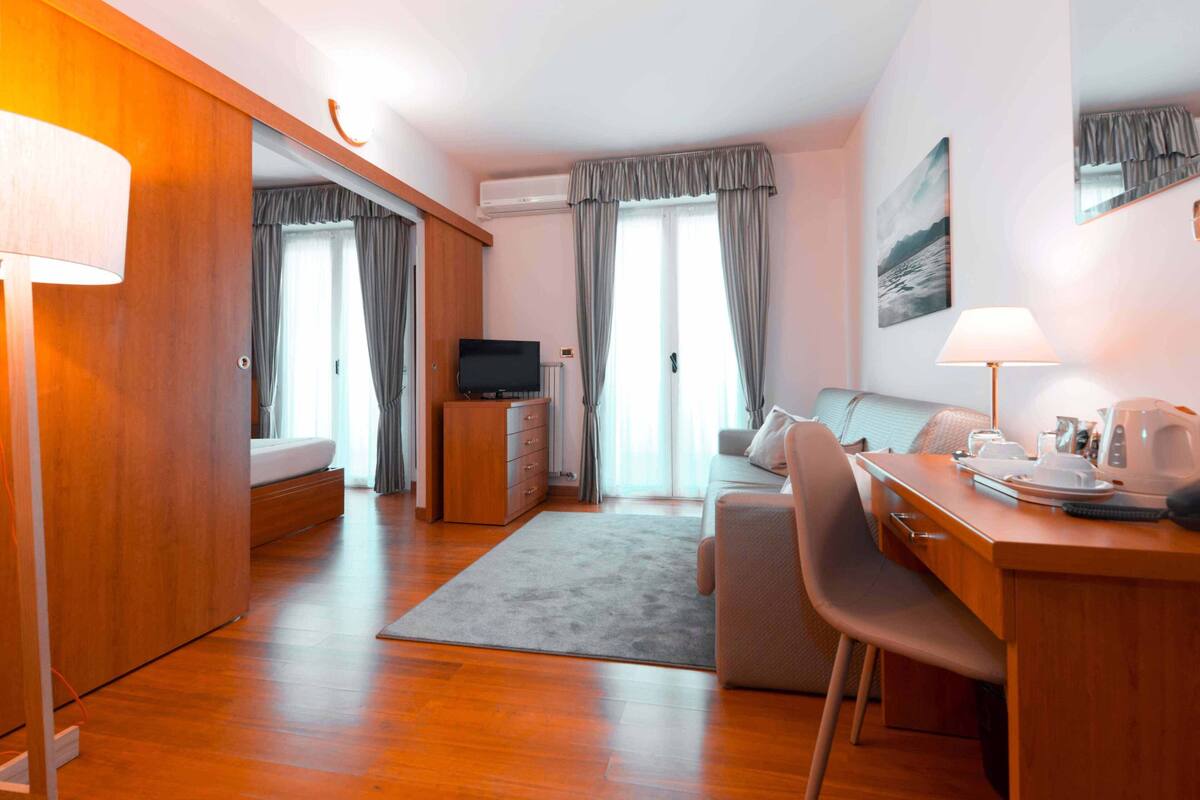 A modern hotel room with a wooden floor, a bed, a desk with a chair, a television, and a dining area.