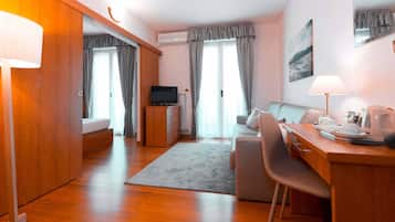 A modern hotel room with a wooden floor, a bed, a desk with a chair, a television, and a dining area.