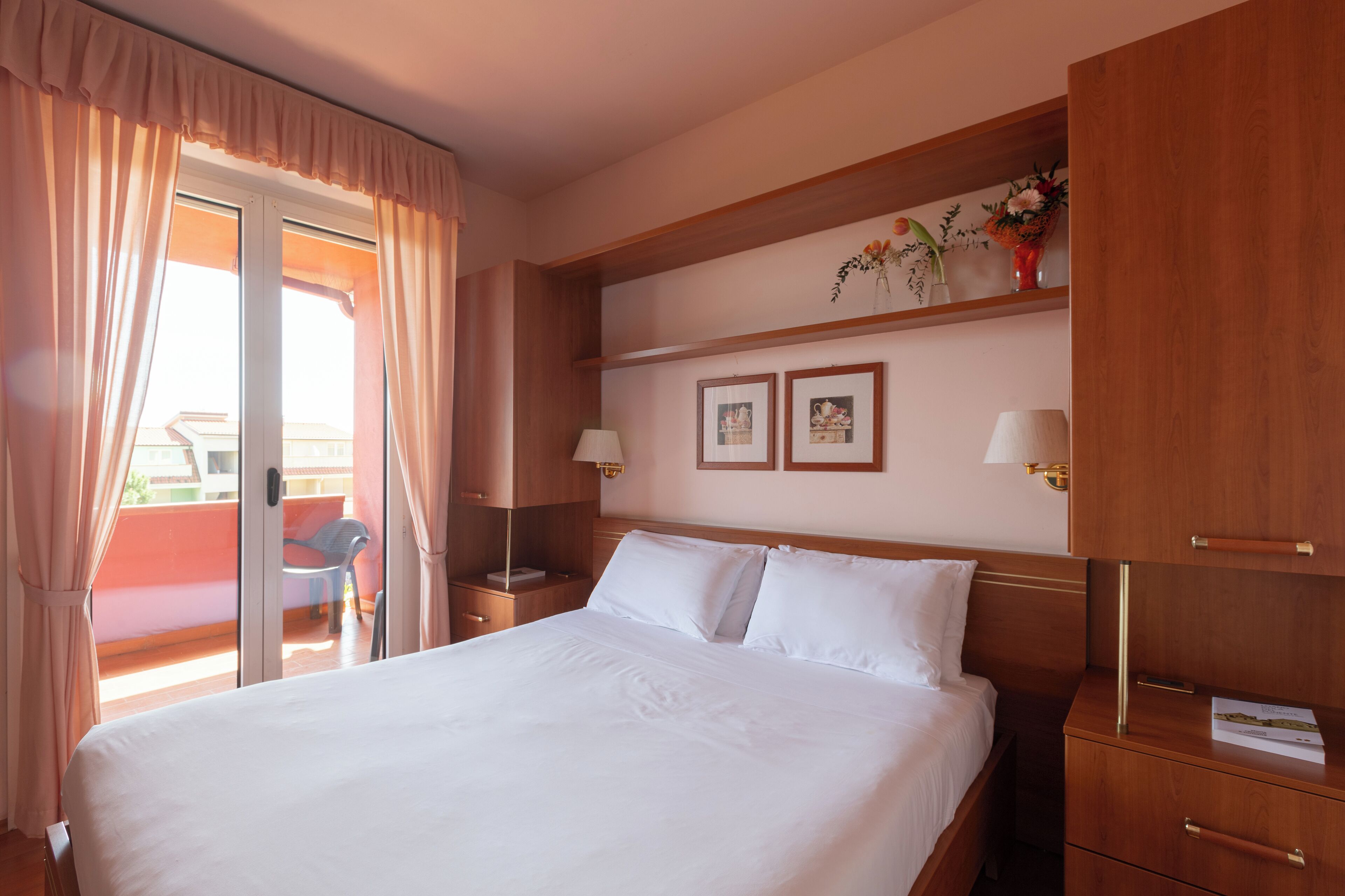 A hotel room with a large bed, wooden headboard, two bedside tables with lamps, a small shelf with framed pictures, and a sliding glass door to the balcony.