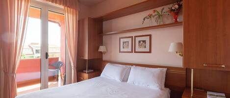 A hotel room with a large bed, wooden headboard, two bedside tables with lamps, a small shelf with framed pictures, and a sliding glass door to the balcony.