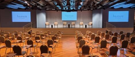 A conference room with rows of chairs facing a stage with a presentation screen displaying "loano2 Village".