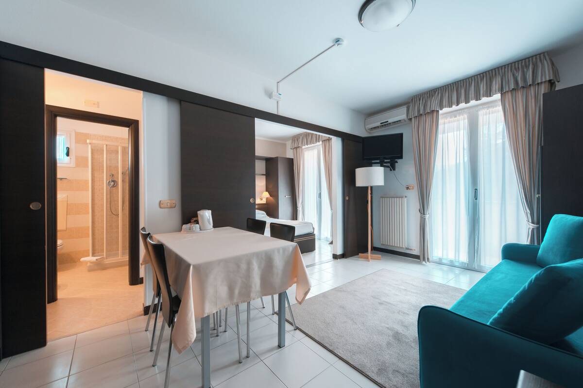 A modern hotel room with a dining area, a sofa, a TV, and a bathroom visible through an open door.