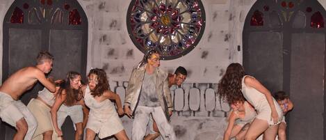 A stage with performers in costumes, a large stained glass window, and stone walls.