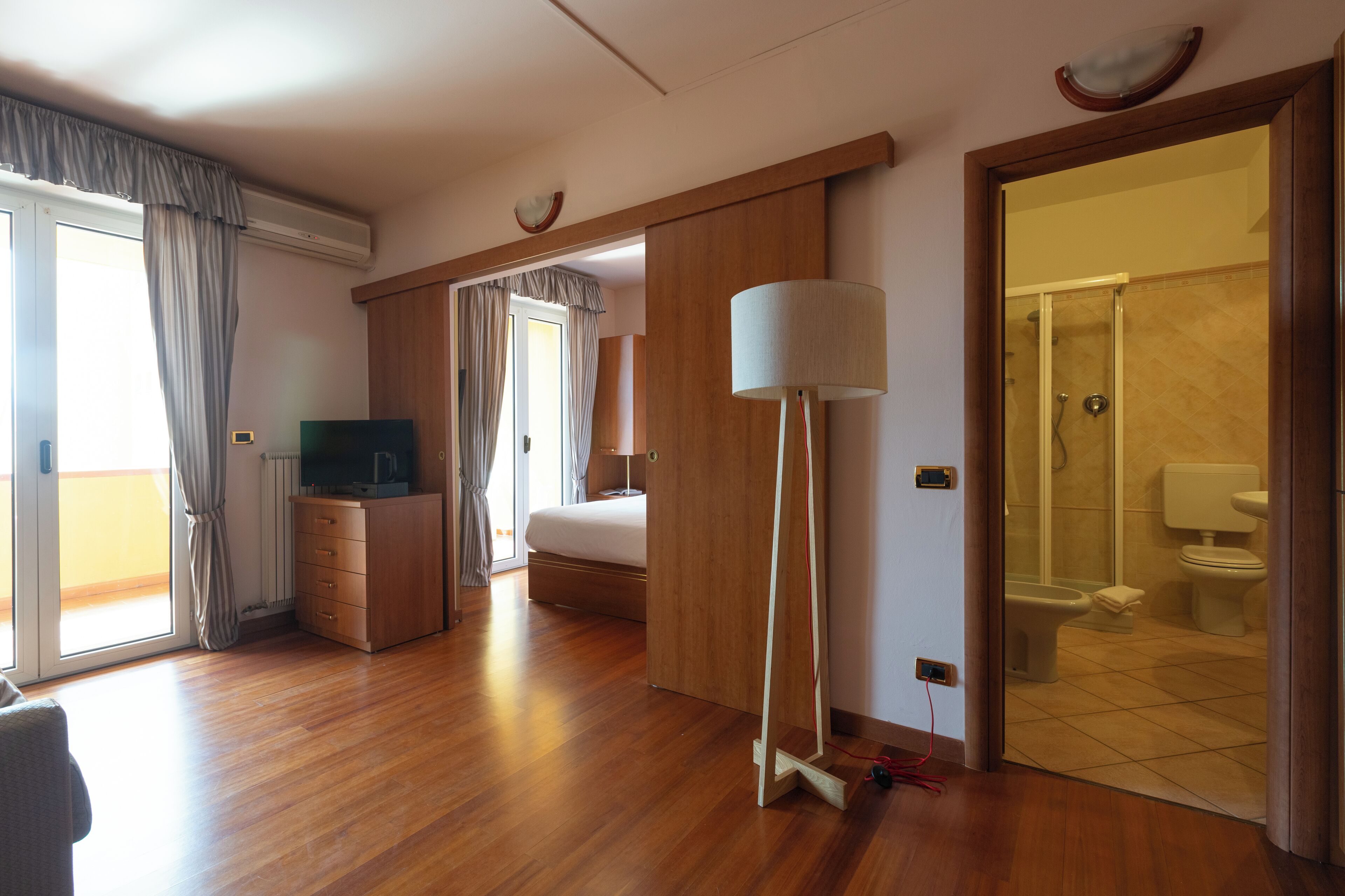 Ein Hotelzimmer mit Holzboden, einem Bett, einem Schreibtisch mit Lampe, einem Fernseher und einem Badezimmer mit Dusche und Toilette.