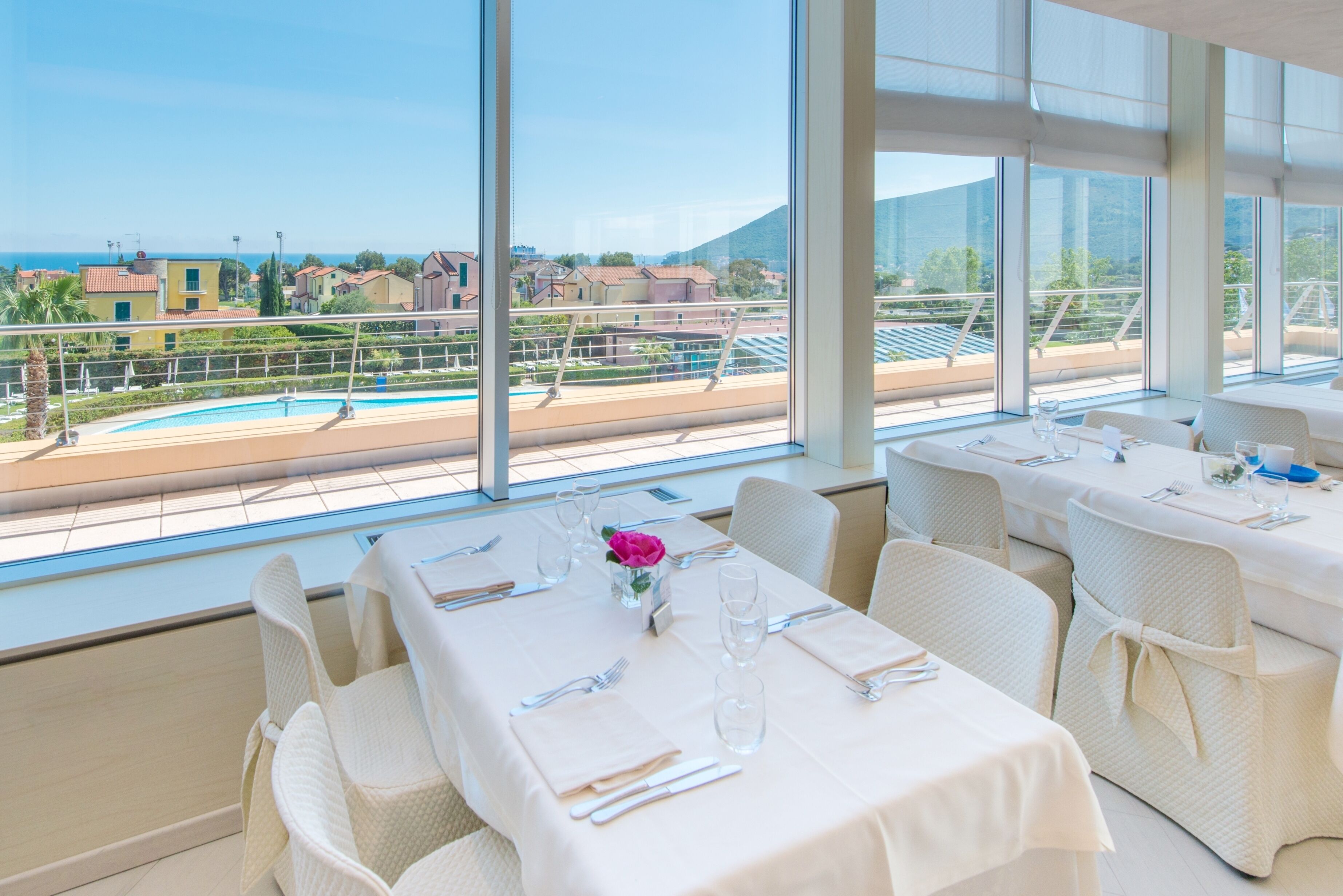 A restaurant with tables set for dining, large windows offering a view of a swimming pool and buildings.