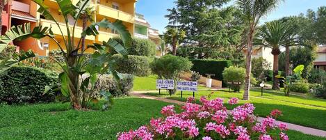 A hotel with a yellow facade, a sign indicating a kids club, and a well-maintained garden with pink flowers.