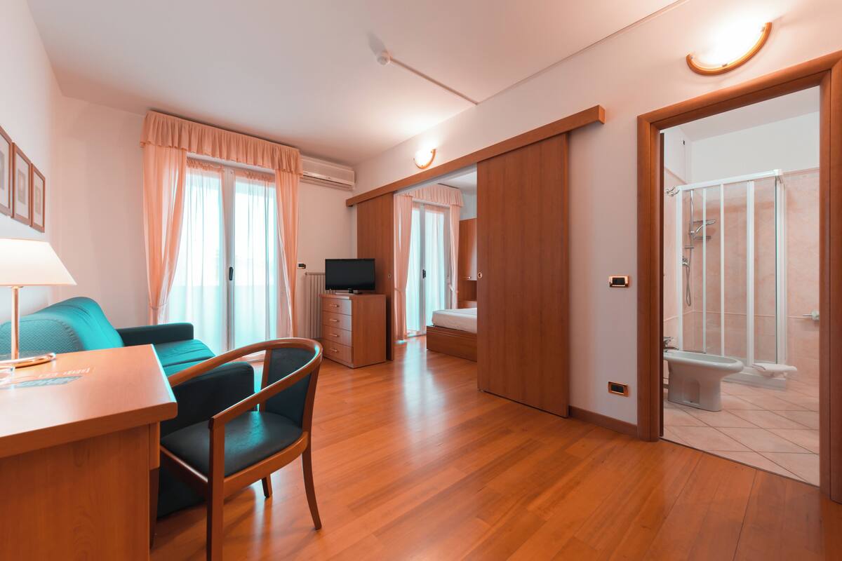 A hotel room with a wooden floor, a sofa, a desk with a lamp, a television, a wardrobe, and a bathroom with a shower and toilet.