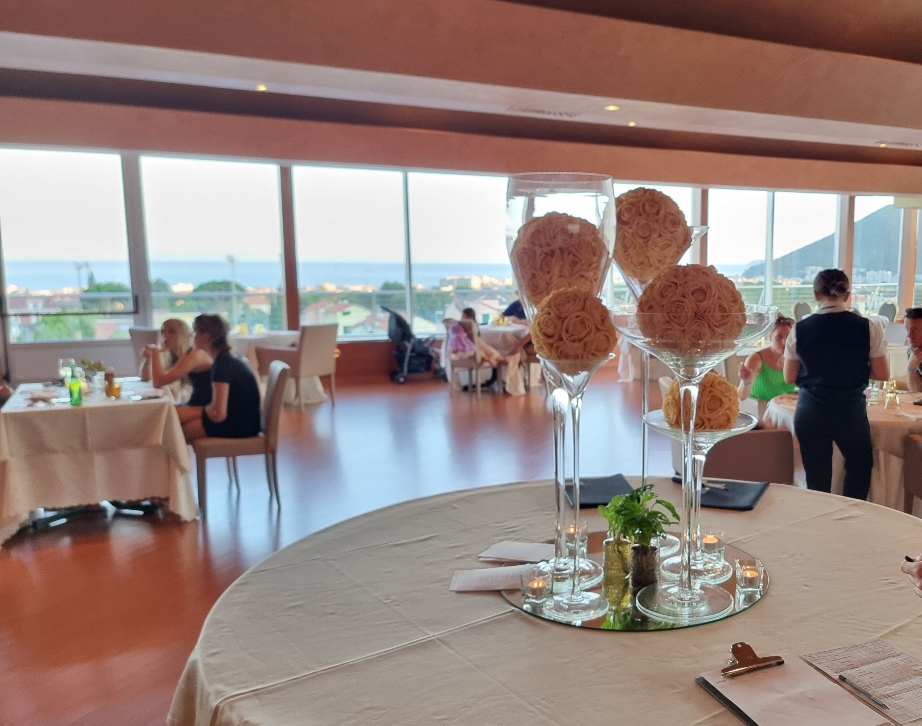 A restaurant with round tables, a view of the sea, and a waiter serving guests.