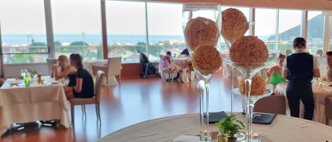 A restaurant with round tables, a view of the sea, and a waiter serving guests.