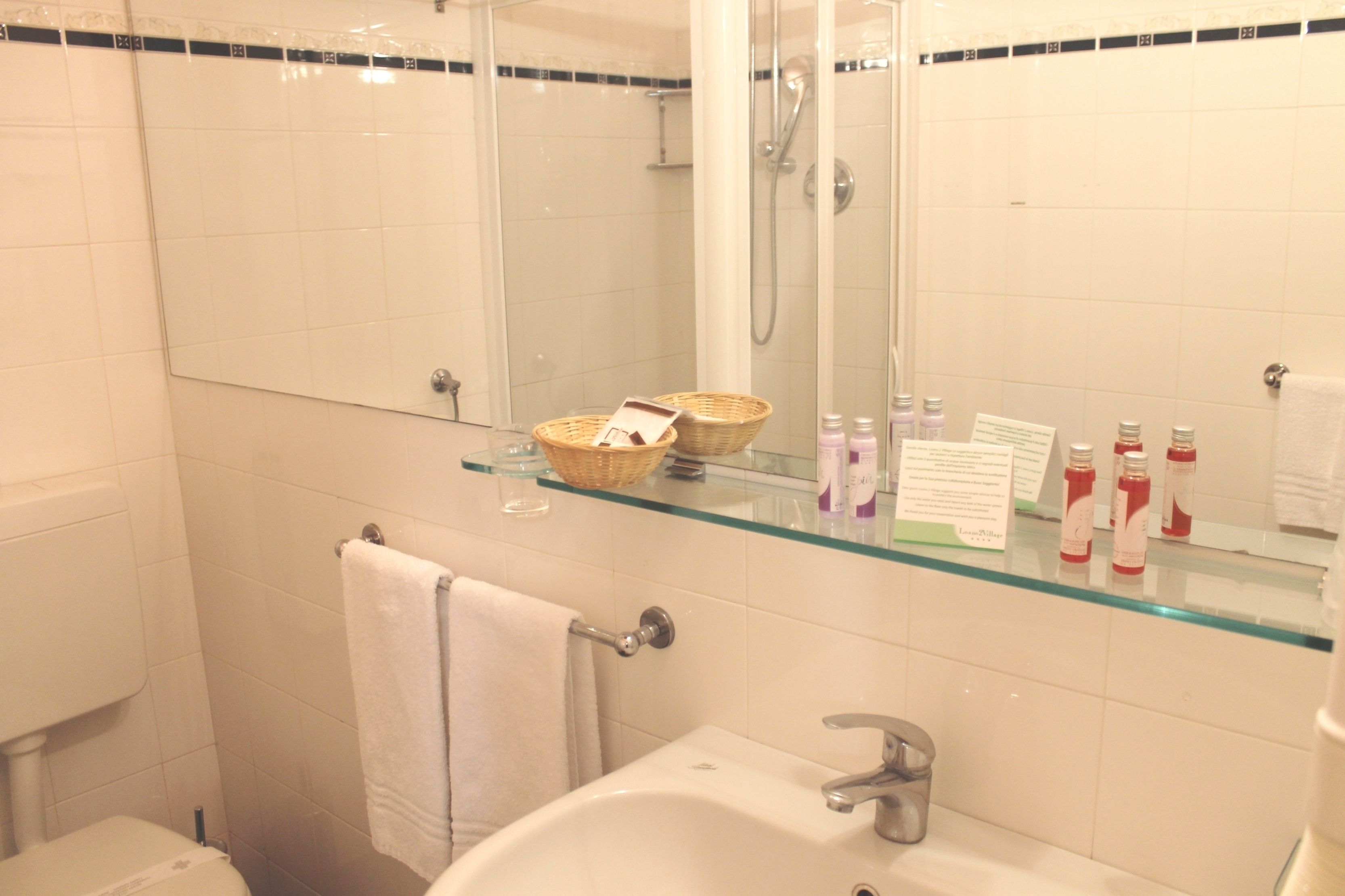 A bathroom with a glass shelf holding toiletries, a white sink, and a glass shower enclosure.