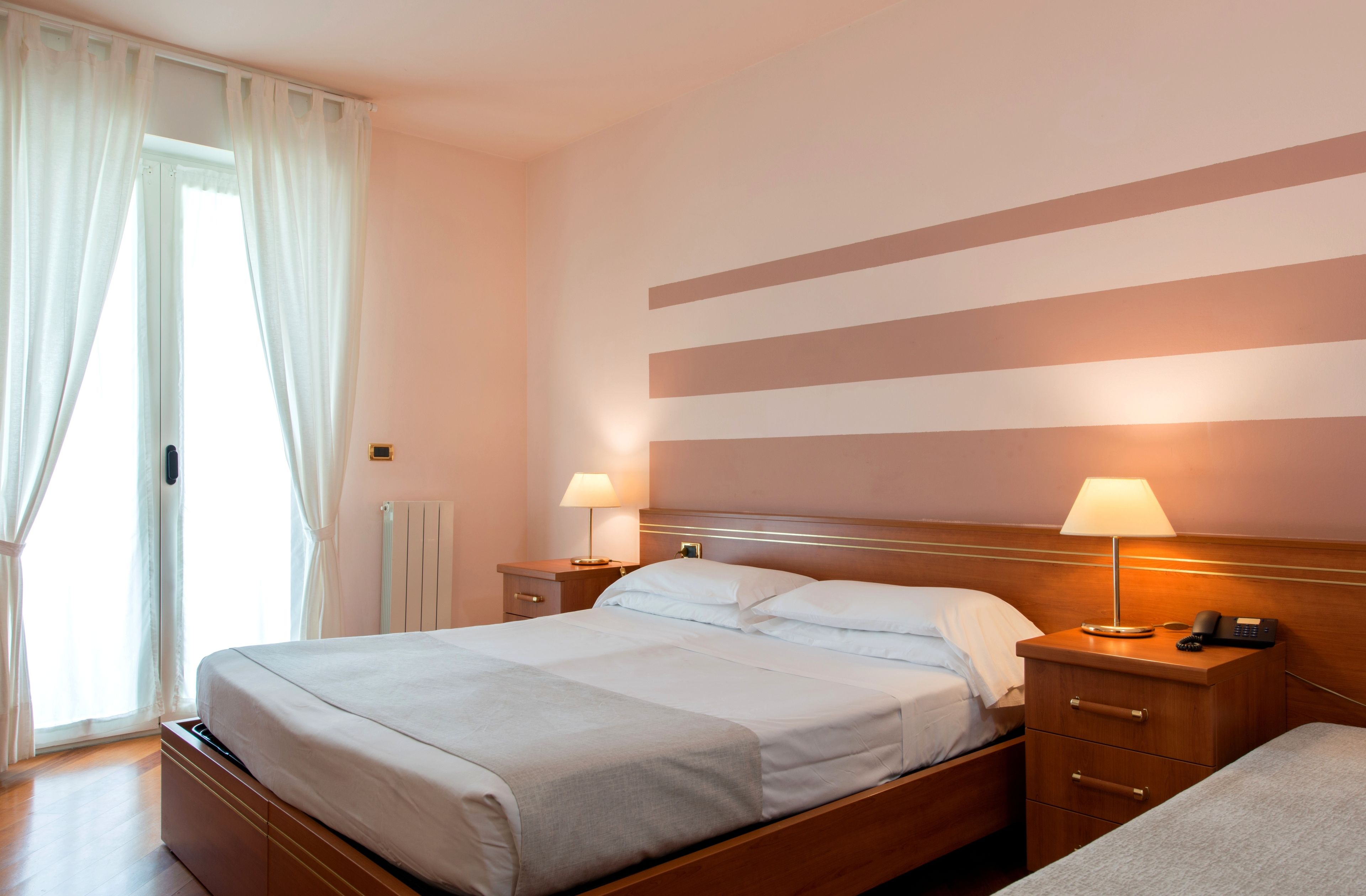 Un cuarto de hotel con una cama grande, dos mesitas de noche con lámparas, un cabecero de madera y vistas al exterior a través de una ventana con cortinas blancas.