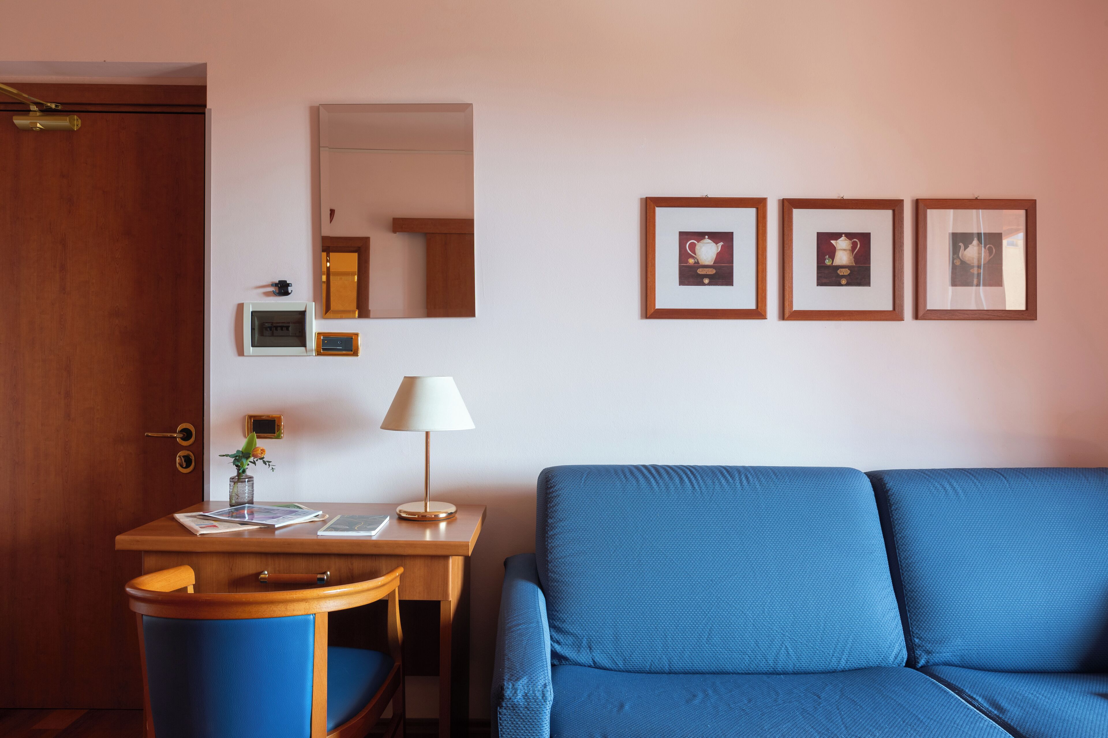 Una habitación con un escritorio de madera, una silla azul, un sofá azul, una lámpara y tres cuadros en la pared.