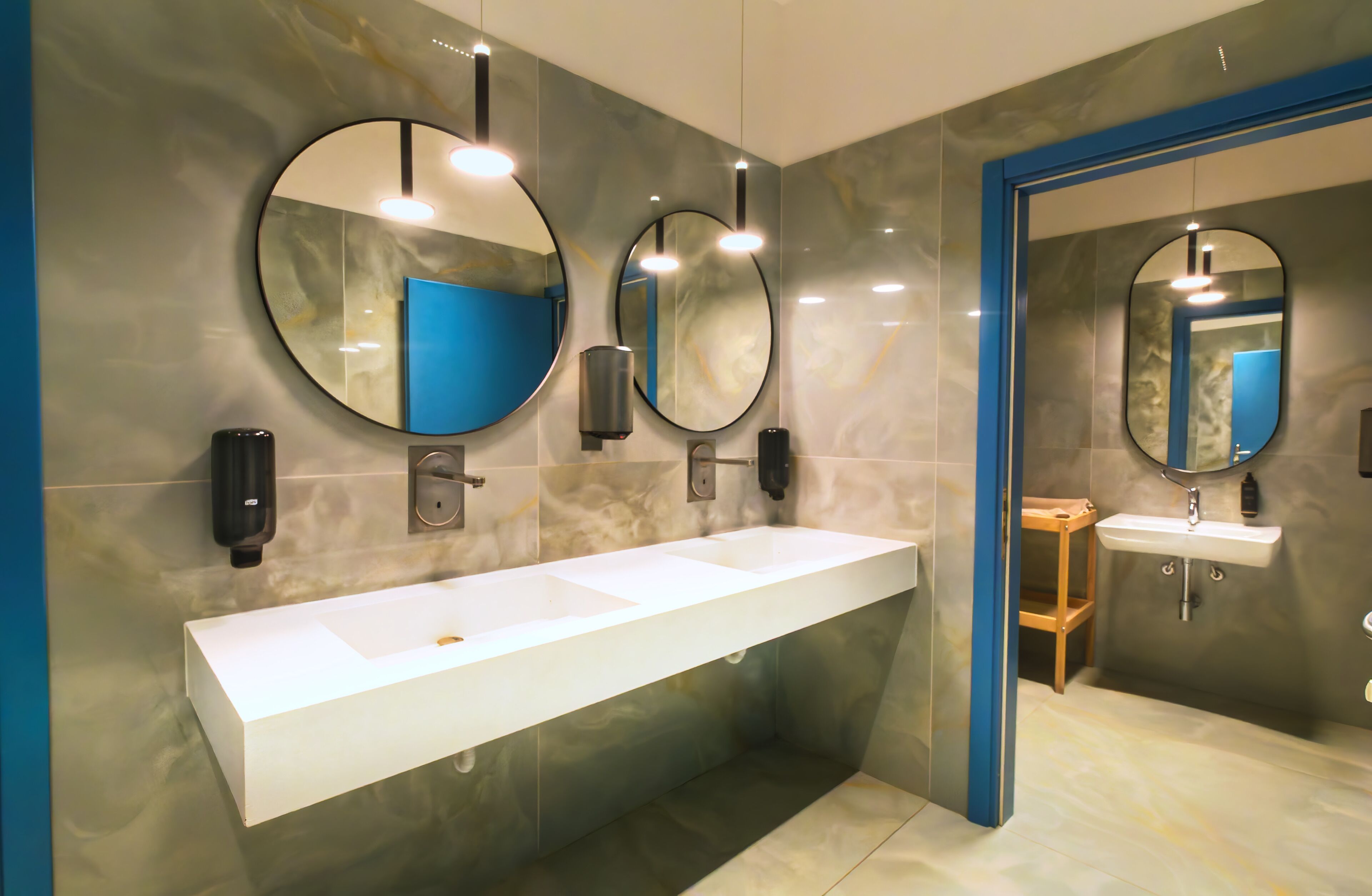 Un baño moderno con un lavamanos blanco grande, dos espejos fijados a la pared y una puerta con marco azul.