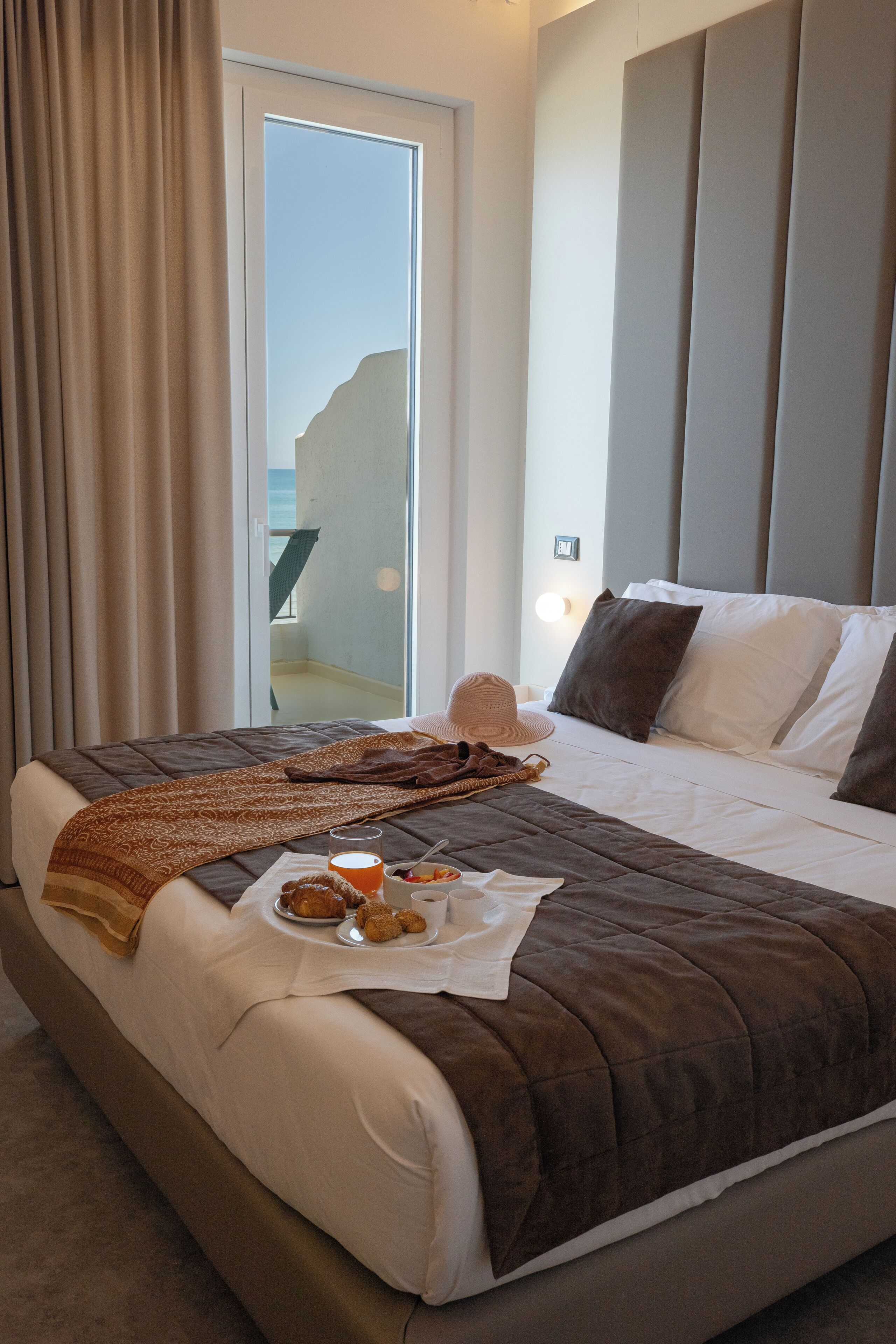 Habitación de hotel con cama, bandeja de desayuno, un sombrero y vista al mar a través de una ventana.