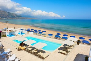 A pool area with multiple lounge chairs and umbrellas, a clear view of the beach, and a mountain in the background. - Hotel La Battigia (Alcamo)