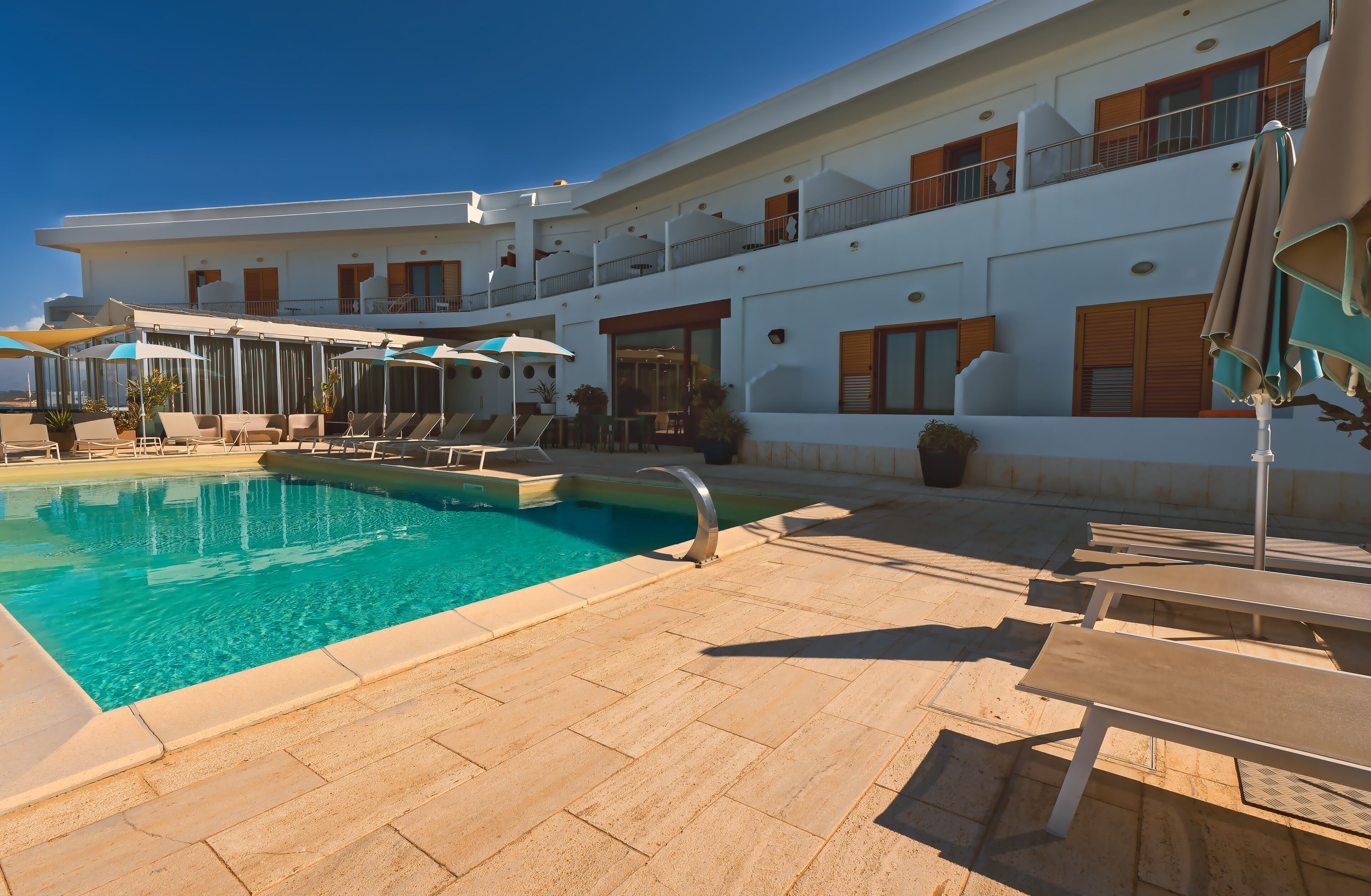 A pool area with lounge chairs and umbrellas, a clear blue pool, and a white building with balconies.