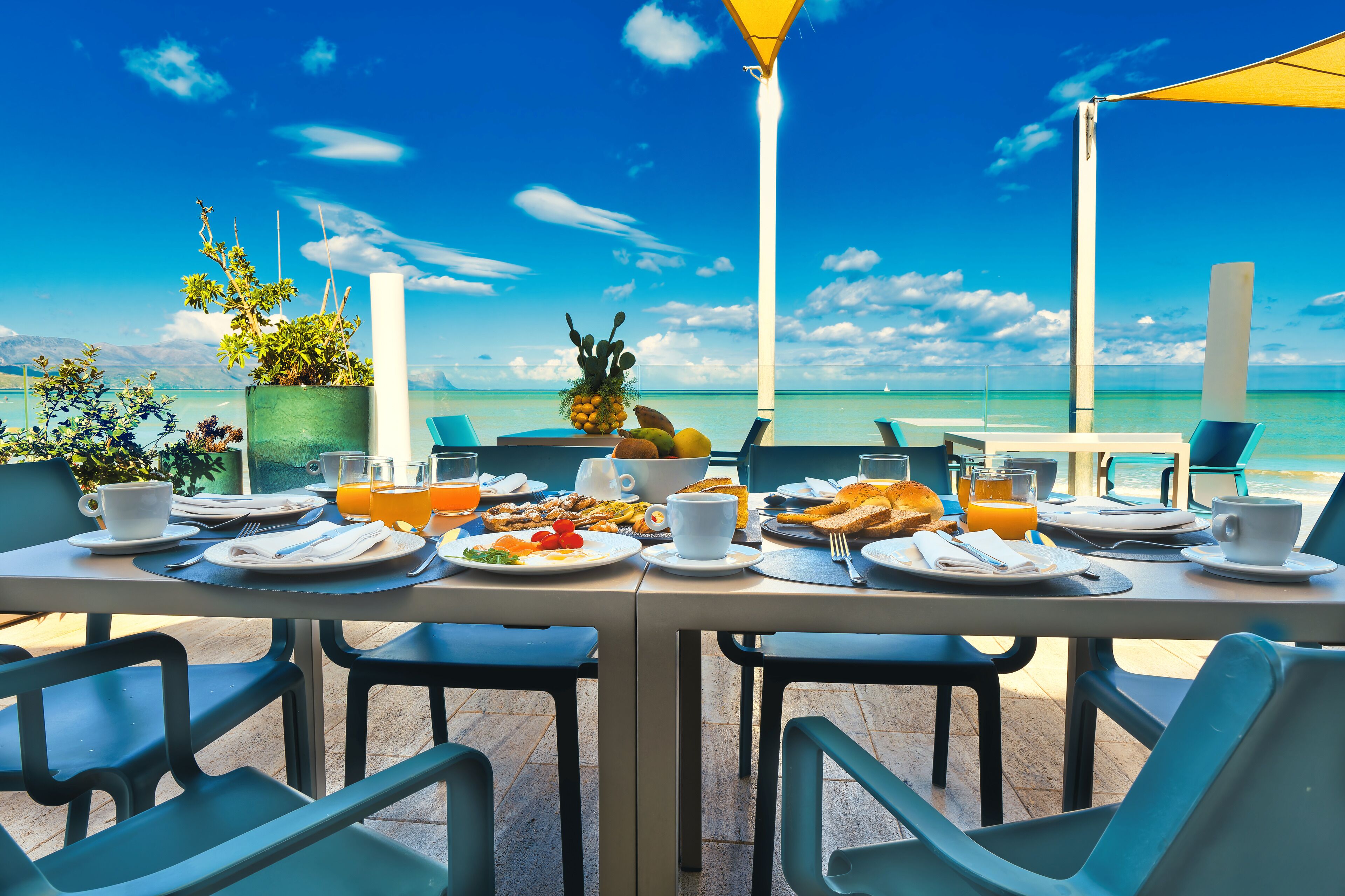 Mesa de desayuno lista con platos, tazas y frutero, con vistas al mar azul y despejado.