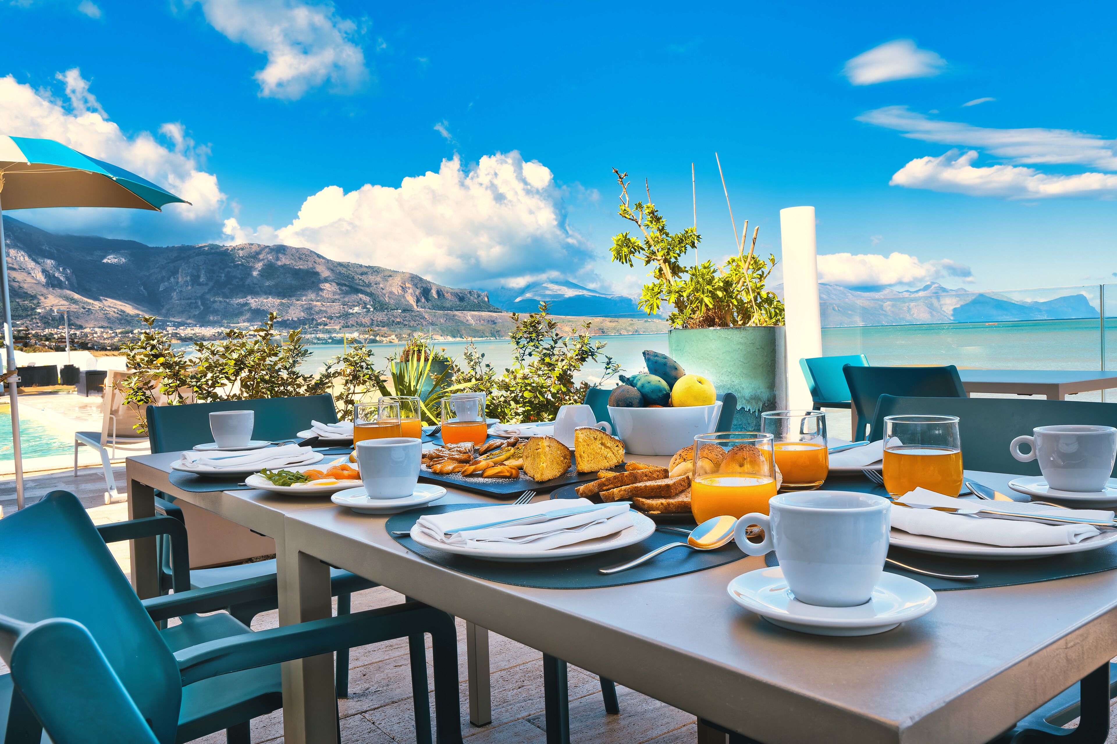 A breakfast table set with plates, cups, and a glass of orange juice, overlooking a scenic lake and mountains.