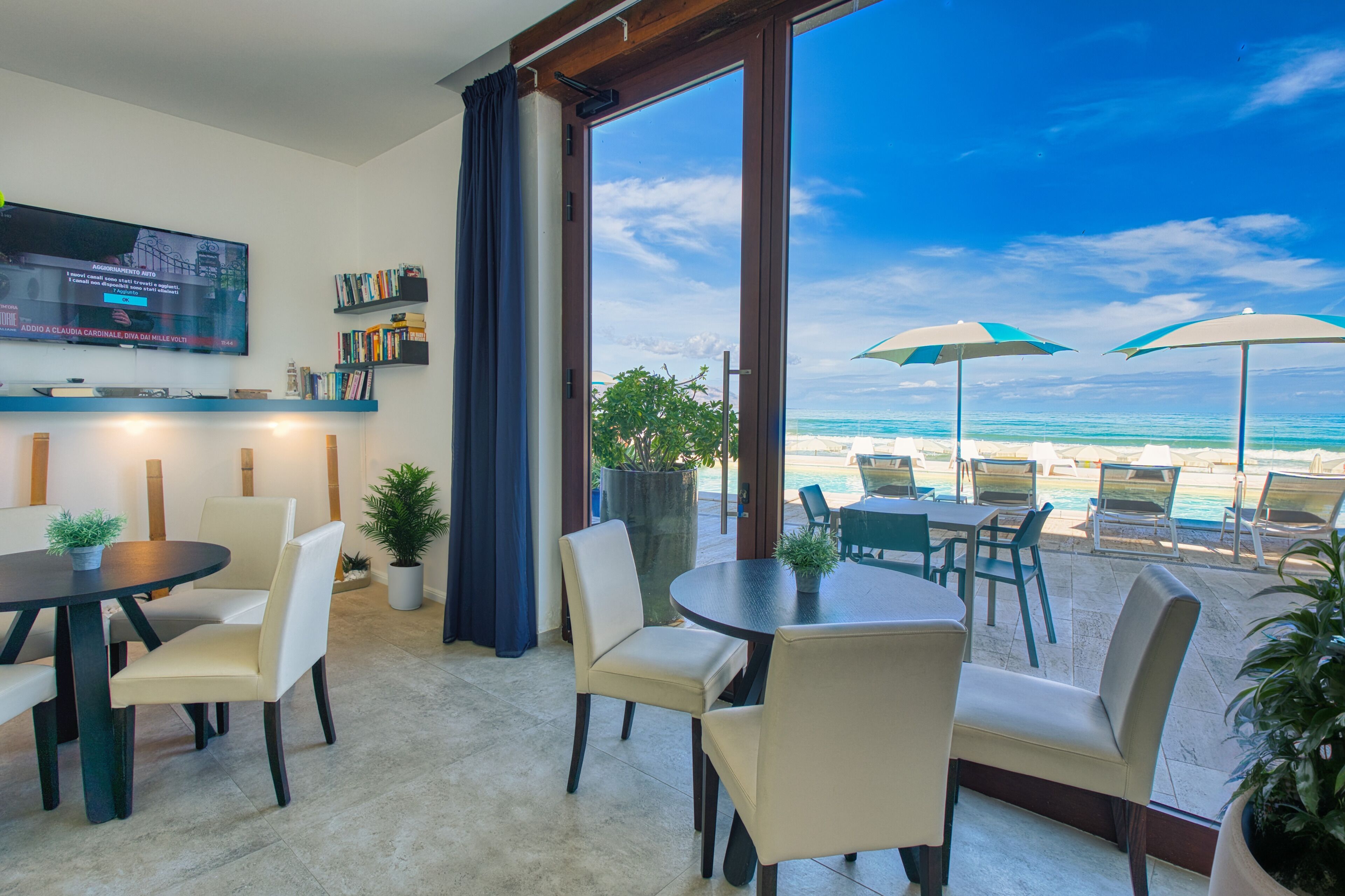 Una habitación con televisor, estanterías, un amplio ventanal con vista a la playa y mobiliario exterior con sombrillas.