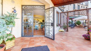An outdoor patio area with tiled flooring, wooden furniture, and potted plants. - Hotel San Trano (Luogosanto)