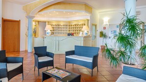 A lobby with wicker furniture, a reception desk, and a large potted plant. - Hotel San Trano (Luogosanto)