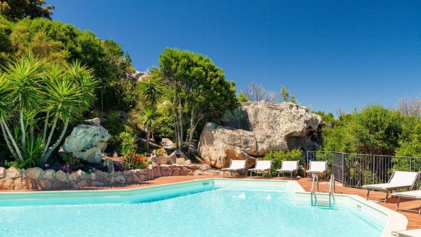 A pool with clear blue water, surrounded by lounge chairs and a stone wall. - Hotel San Trano (Luogosanto)