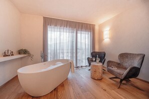 A modern bathroom with a freestanding bathtub, a wooden stool, and a seating area with a chair and a table. - Cristallo Levico Terme (Levico Terme)