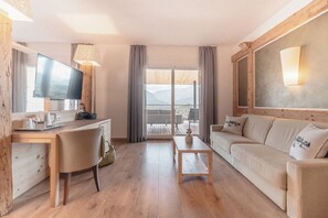A hotel room with a wooden desk, a television, a sofa, and a balcony with a view of mountains. - Cristallo Levico Terme (Levico Terme)