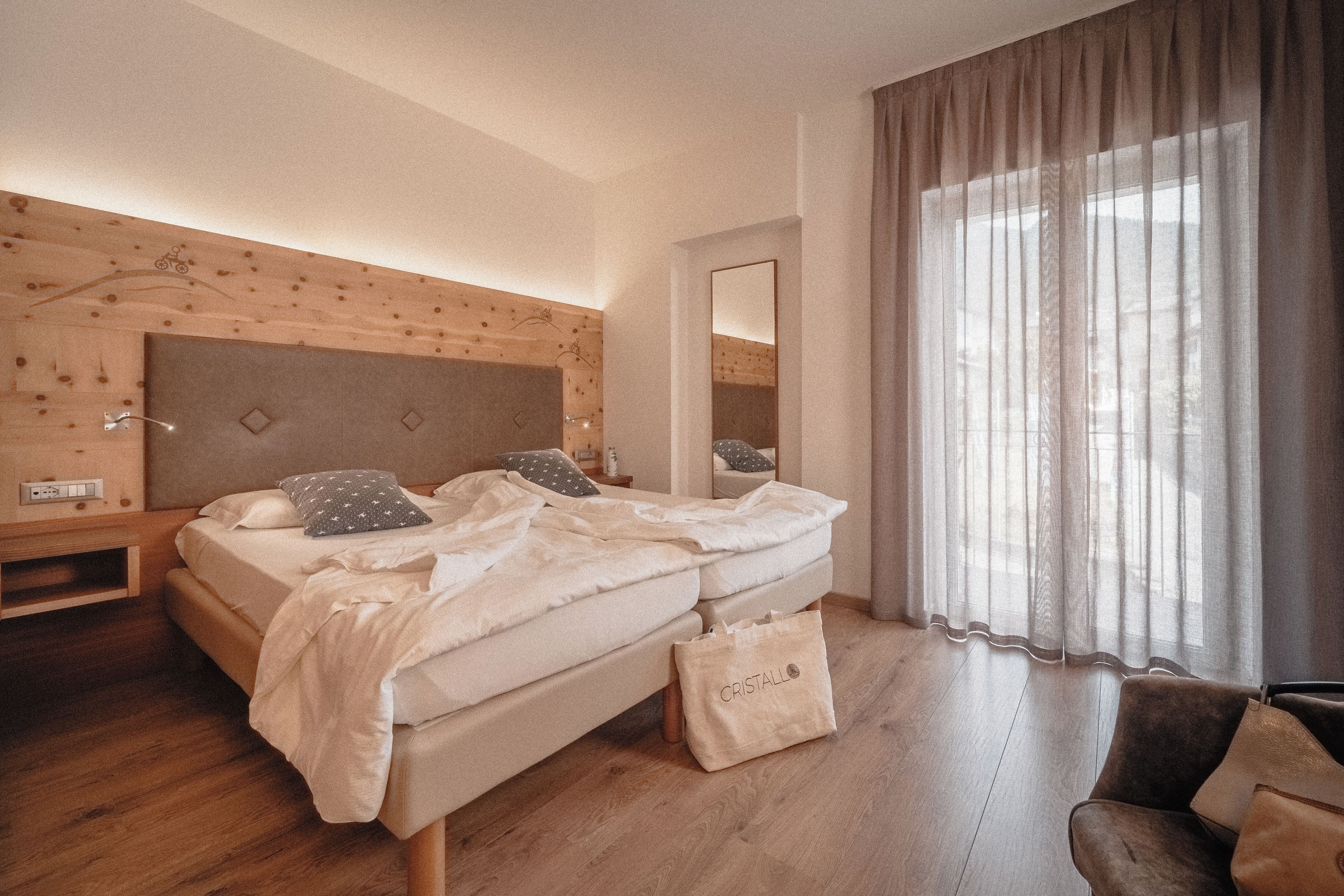 A bedroom with a wooden headboard, a bed with pillows, a bedside table, a mirror, a window with curtains, and a bag on the floor.