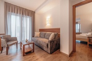 A hotel room with a wooden floor, a sofa, a chair, a small table, and a bed with a wooden headboard. - Cristallo Levico Terme (Levico Terme)