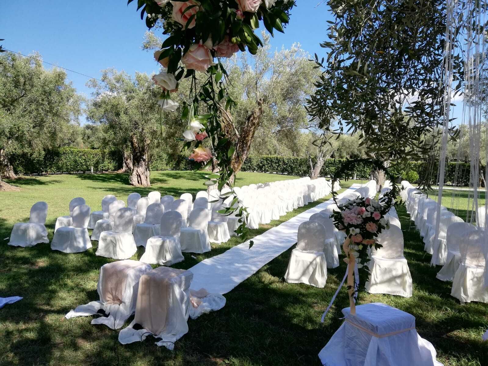 An outdoor wedding setup with rows of chairs and tables covered in white cloths, a floral arrangement, and a white aisle runner.