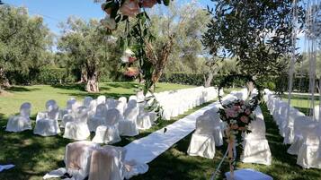 Montaje para boda al aire libre con filas de sillas y mesas cubiertas con manteles blancos, un arreglo floral y una alfombra blanca para el pasillo.