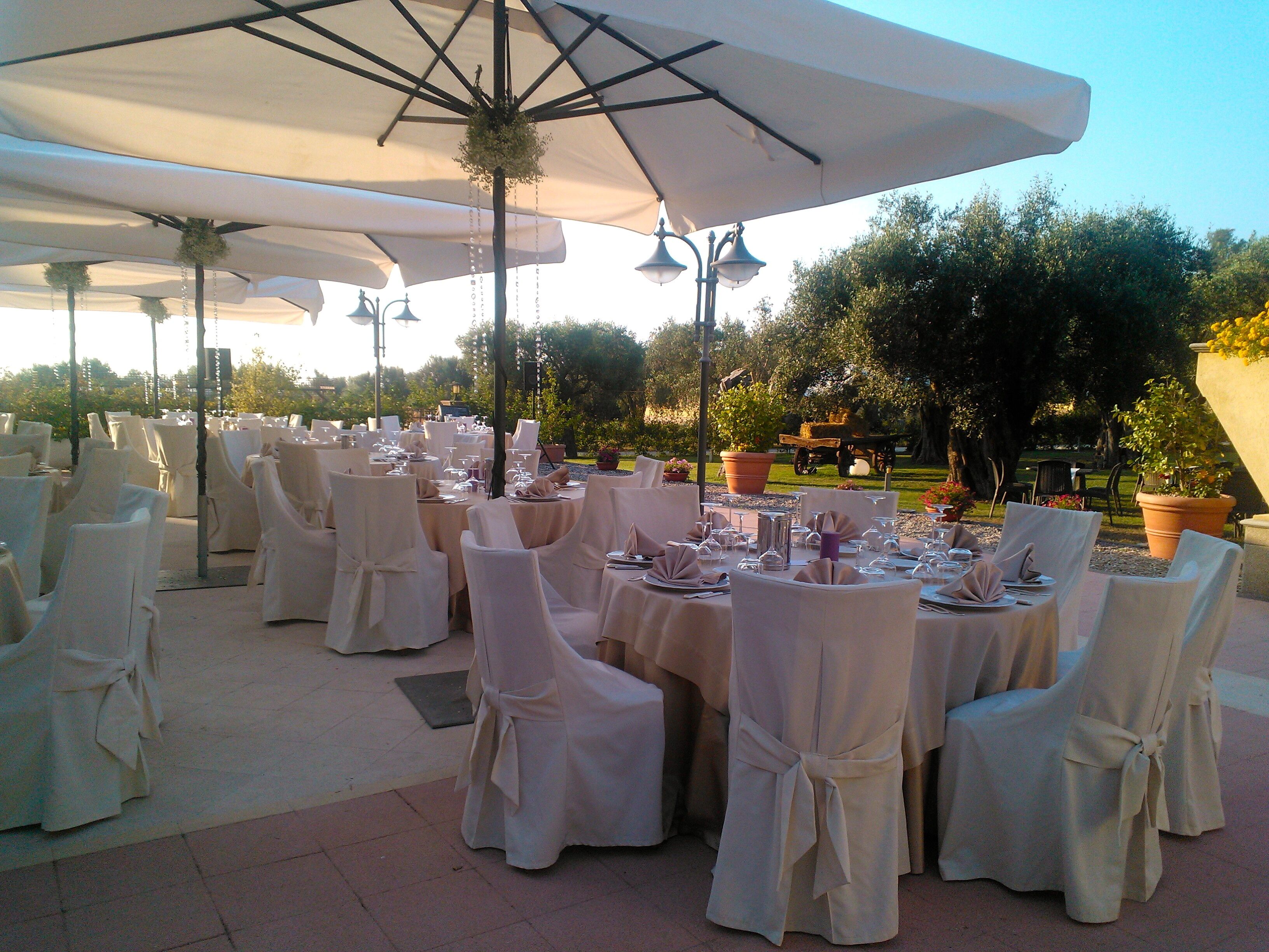 Un área de comedor al aire libre con mesas redondas, sillas blancas y sombrillas grandes.