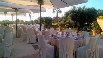 Un área de comedor al aire libre con mesas redondas, sillas blancas y sombrillas grandes.