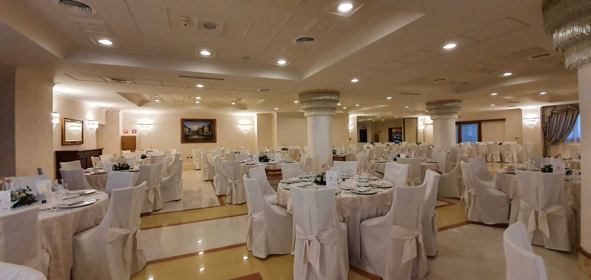 Een balzaal met ronde tafels, witte tafellakens en stoelen, met een hoog plafond, inbouwverlichting en grote zuilen.