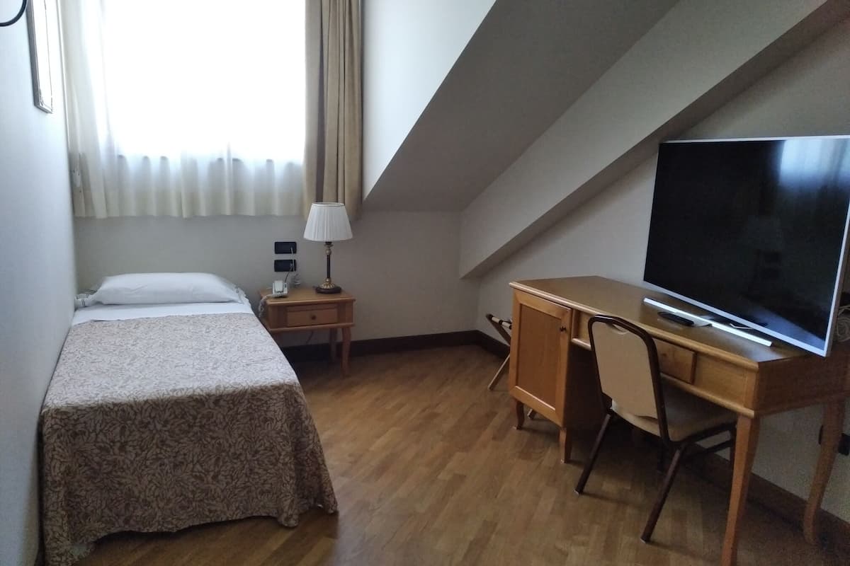 A small, neatly arranged room with a bed, a desk, a chair, a television, and a lamp.