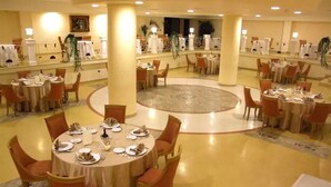 Un espacioso comedor de hotel con varias mesas redondas y sillas, una zona circular decorativa en el centro y vistas del vestíbulo al fondo.