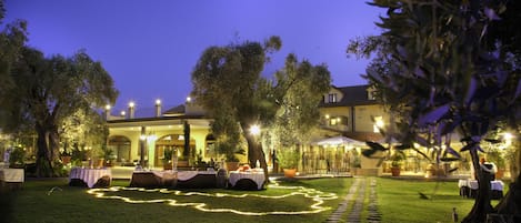 An outdoor dining area with tables set up under trees, a pathway leading to a building, and a well-lit entrance.