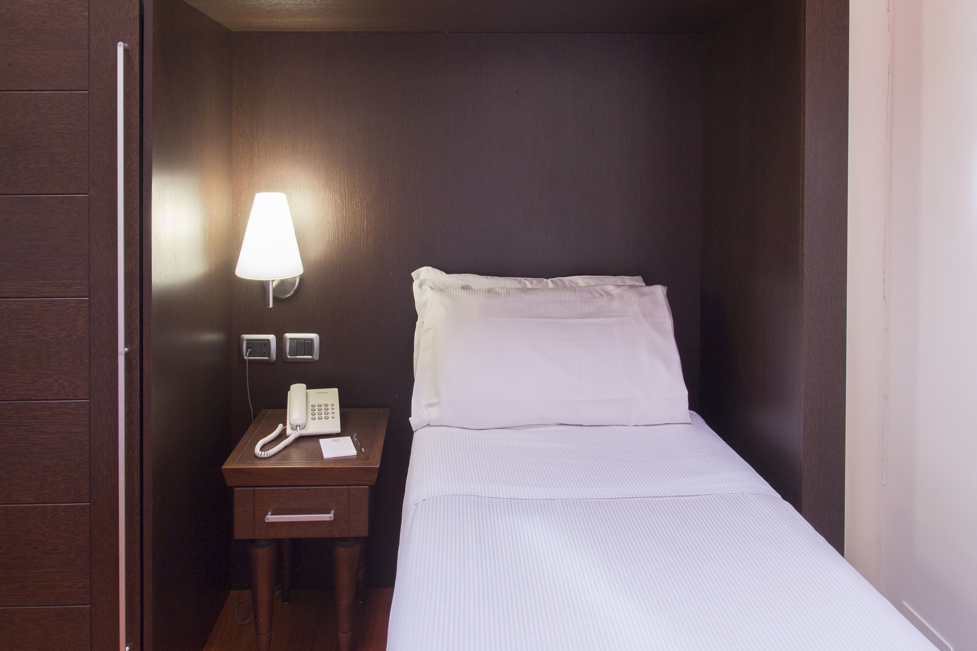 A small, neatly made hotel room with a single bed, a bedside table with a lamp, a phone, and a small drawer.