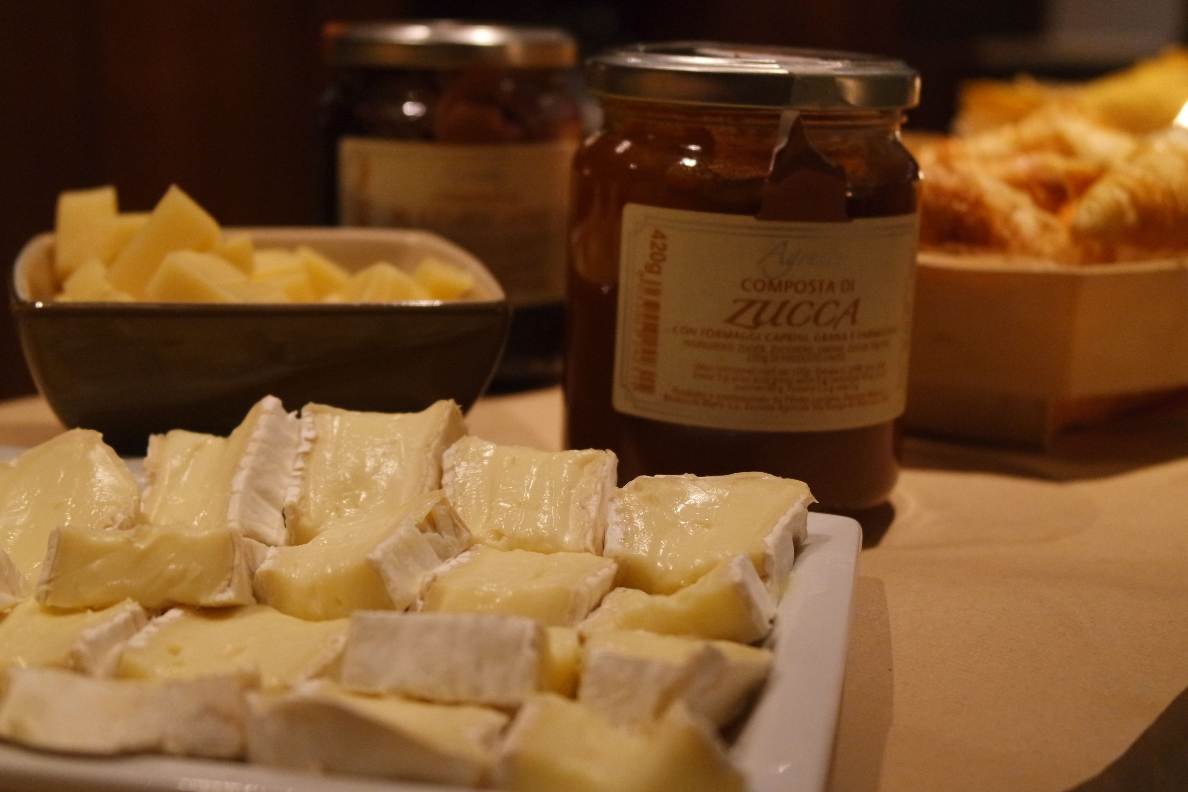 A table with a bowl of cubed cheese, a jar of Zucchini, and a box of bread.