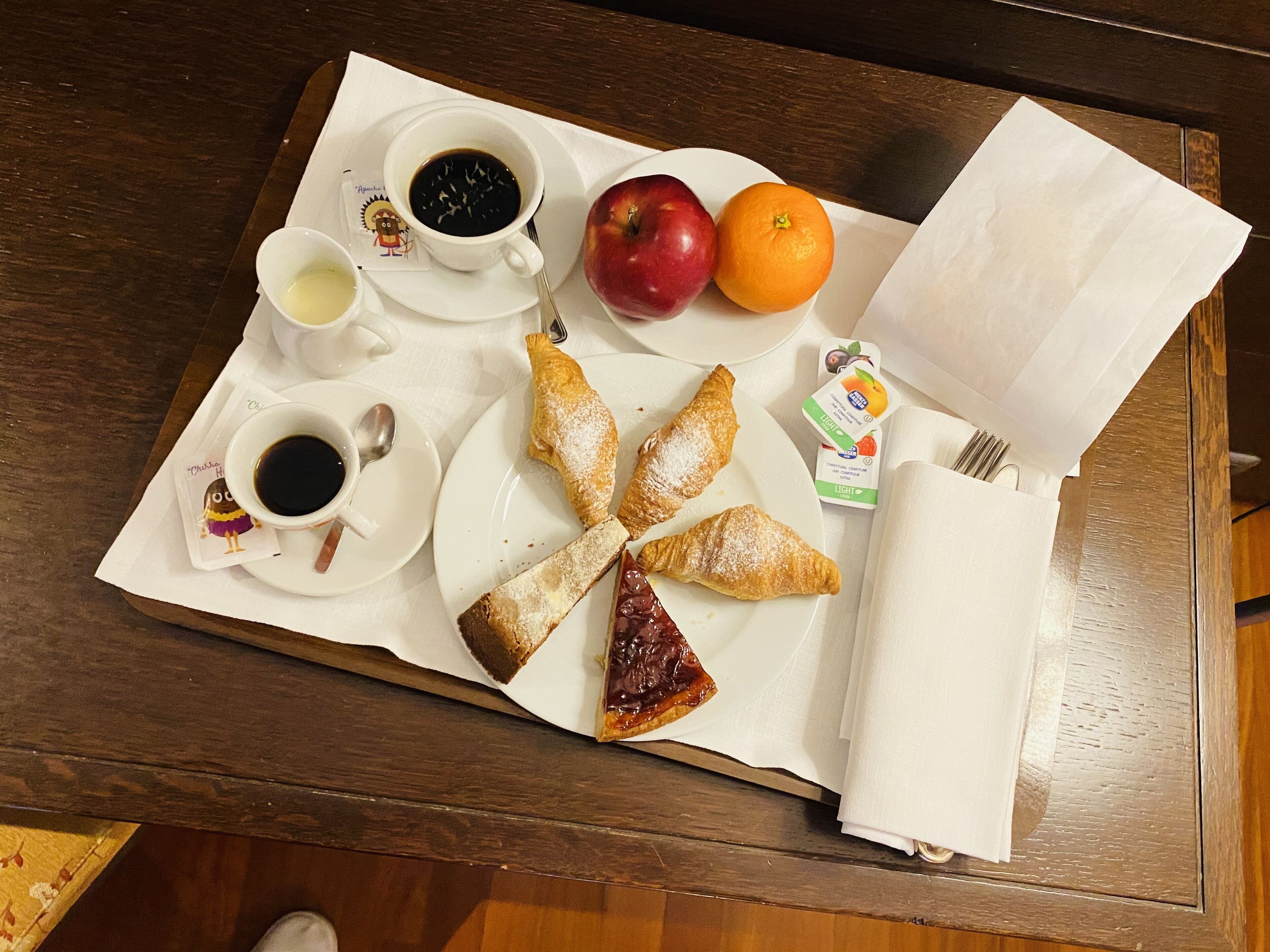 Un plateau de petit-déjeuner avec des croissants, une pomme rouge, une orange et deux tasses de café, posé sur une table en bois.