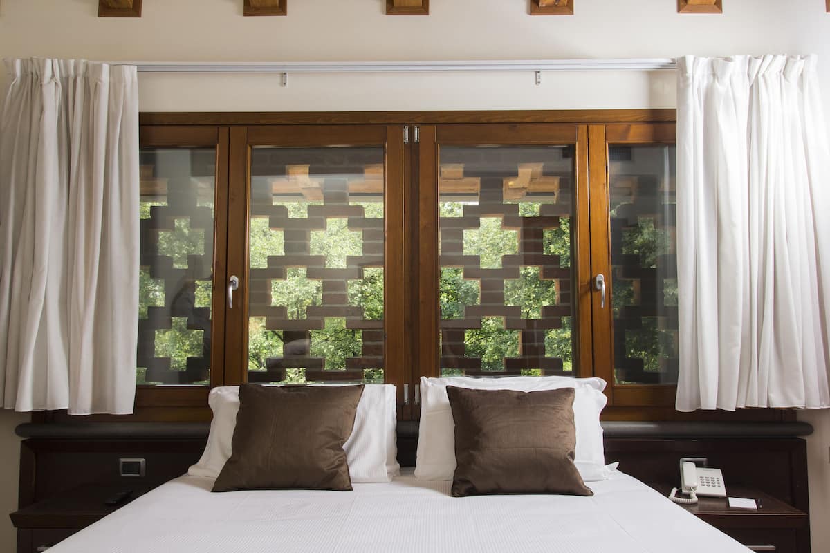 A hotel room with a large bed, two pillows, a bedside table with a phone and a lamp, and a window with curtains.