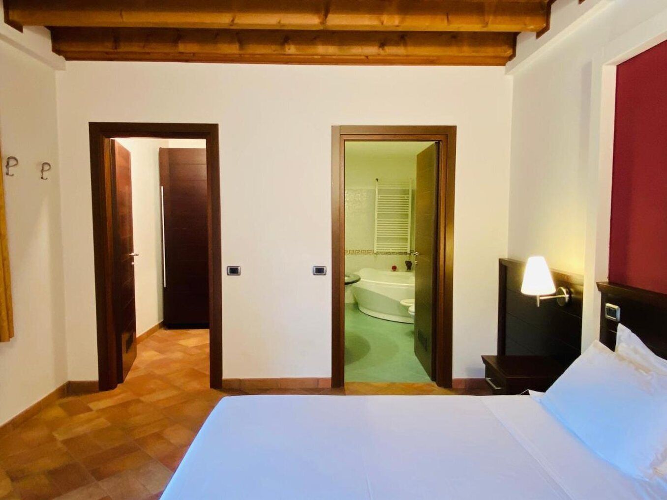 A hotel room with a bed, a nightstand with a lamp, a wooden ceiling, and a view of a bathroom with a bathtub and toilet.