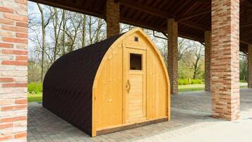 Une cabane en bois avec un toit à deux versants, une petite fenêtre et une porte. Elle est située sous une zone abritée, soutenue par des piliers en briques.
