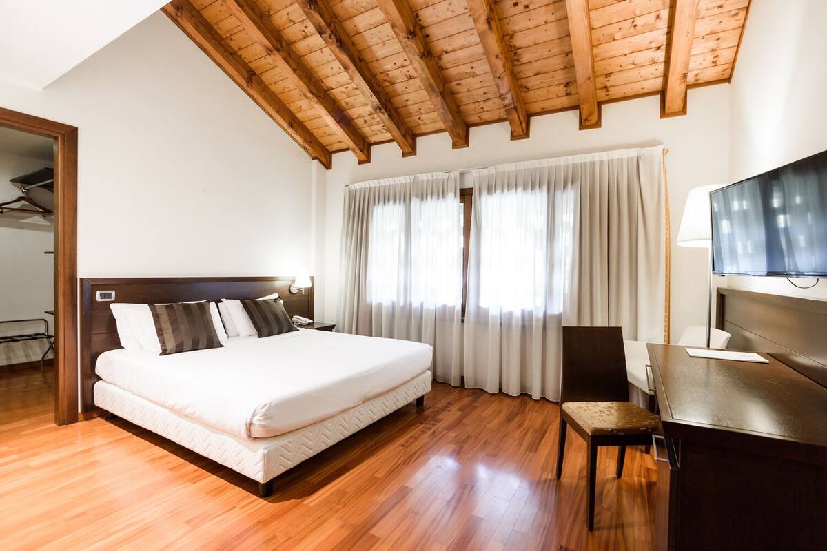 A hotel room with a wooden ceiling, a large bed with a headboard, a desk with a chair, and a TV.