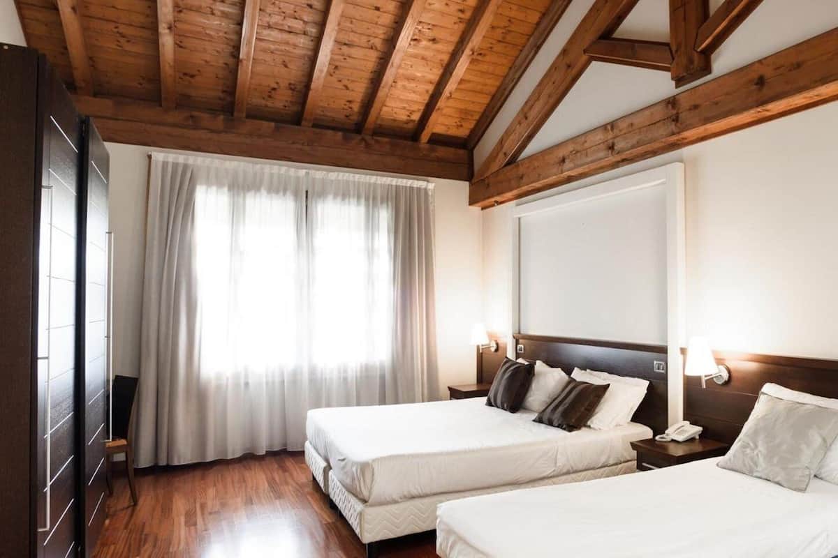 A hotel room with two beds, wooden beams on the ceiling, a large window with curtains, and bedside lamps.