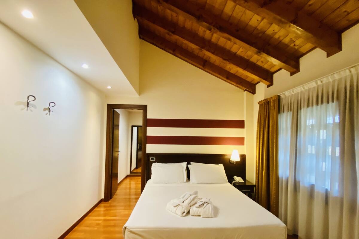 A hotel room with a wooden ceiling, a bed with white linens, a nightstand with a lamp, and a view of a corridor.