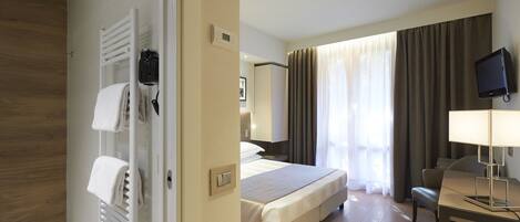 Habitación de hotel con cama, escritorio, televisor y baño con toallero y radiador.