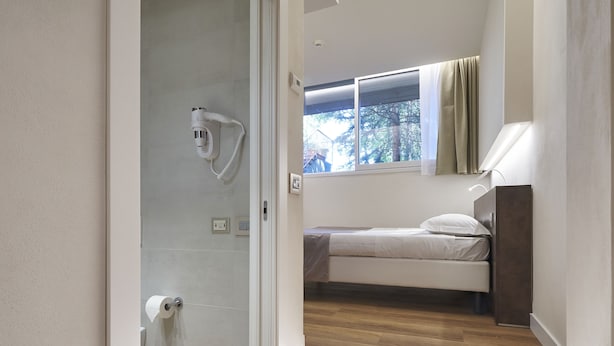 A modern bedroom with a wooden floor, a bed with a headboard, a window with curtains, and a shower area with a towel rack. - Park Hotel Chianti (Barberino Tavarnelle)