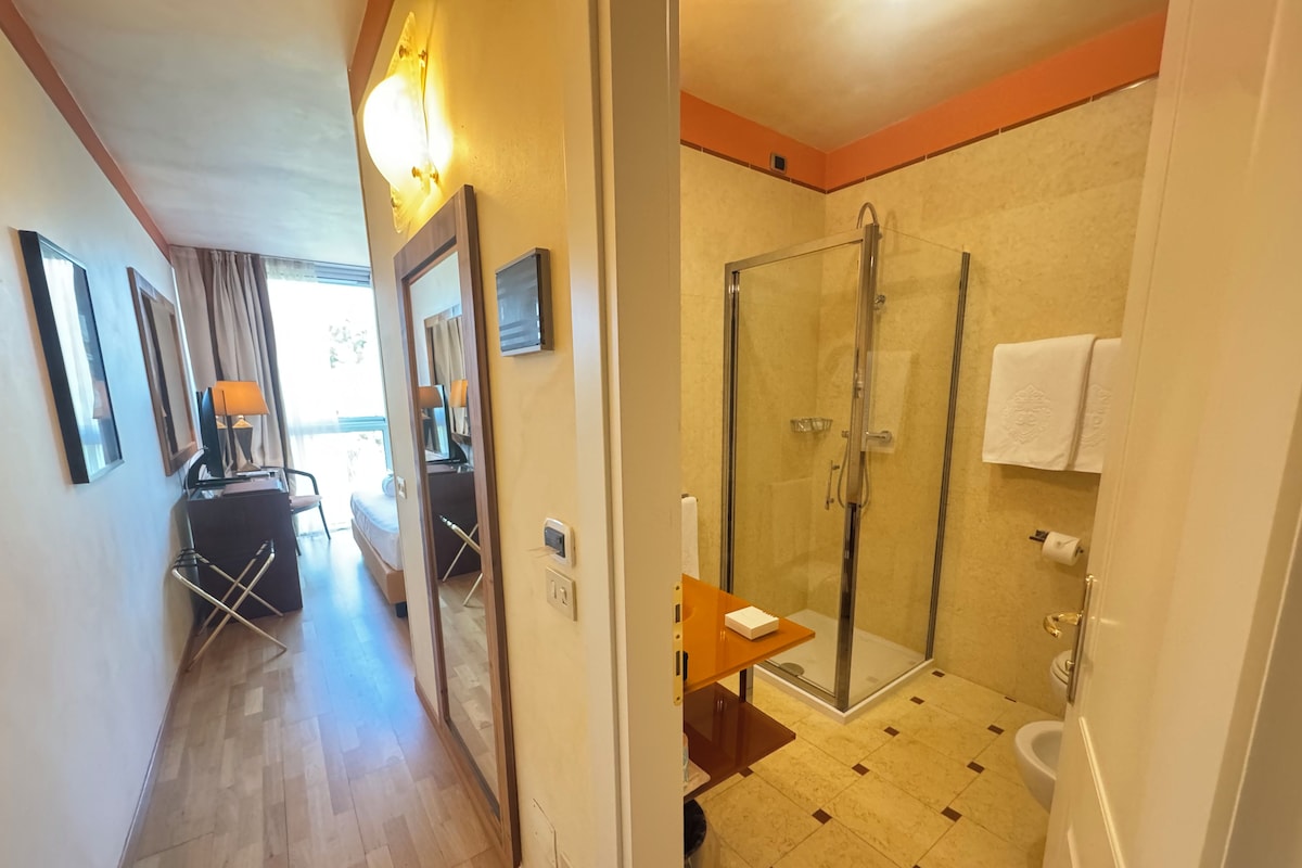 A hotel room with a wooden floor, a desk with a lamp, a bed, a shower with a glass door, and a toilet.